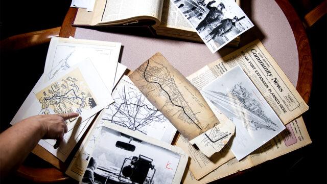 Historical maps, newspapers, photographs and printed volumes spread across a table during a research session.