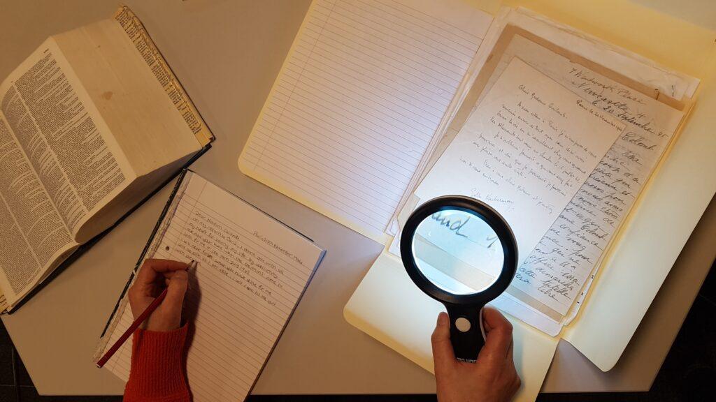 A researcher examining a handwritten nineteenth-century letter with a magnifying glass beside an open reference volume and notebook.