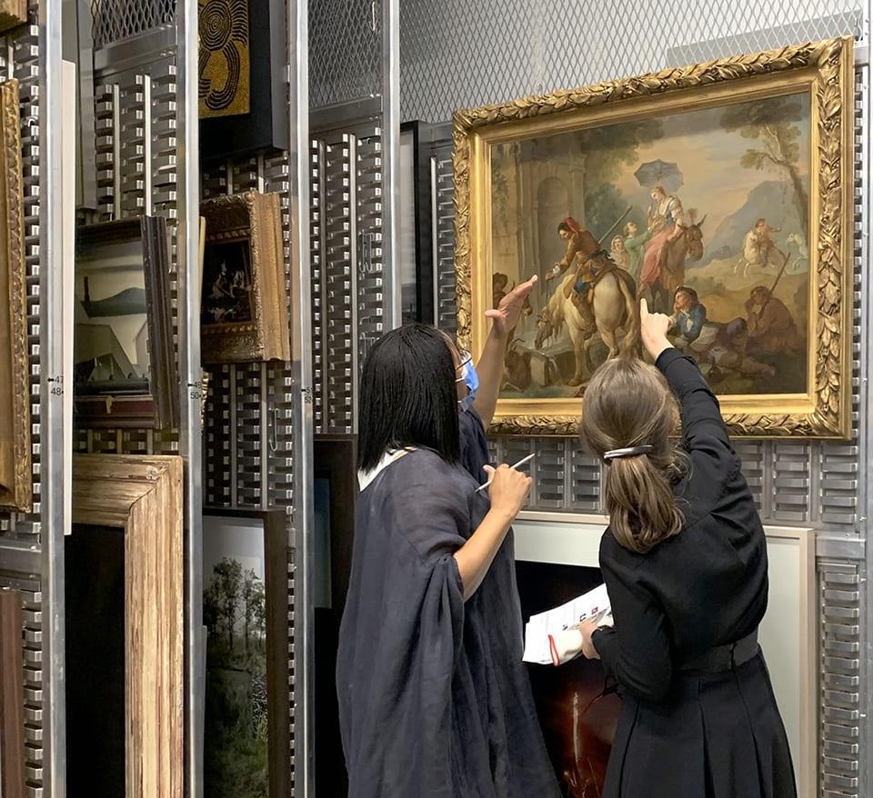 Two curators examining a gilt-framed painting on a sliding rack inside a museum collection store.
