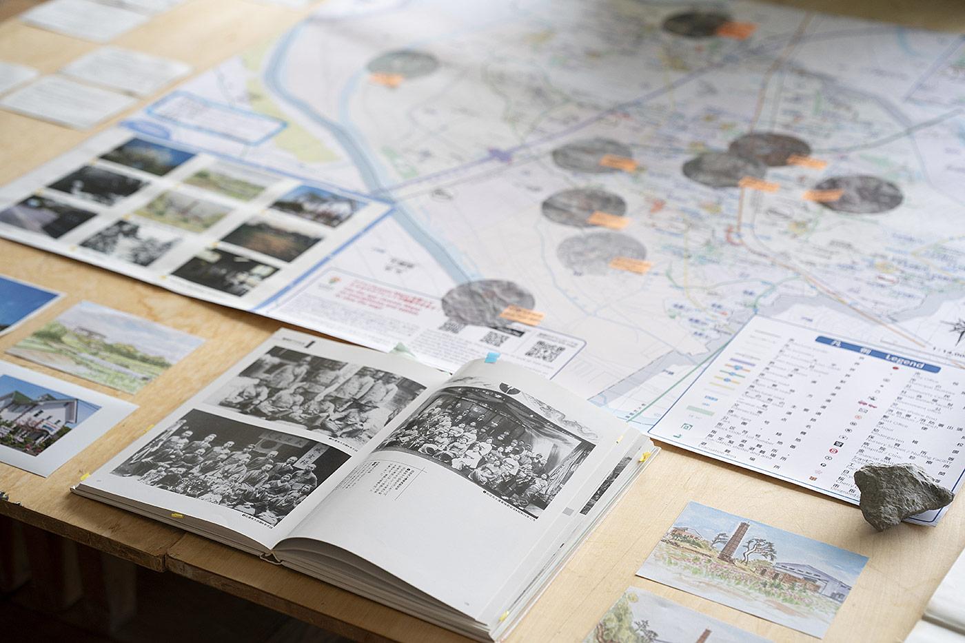 A research table layered with historical maps, an open illustrated volume of archival photographs, site drawings and annotated documents.